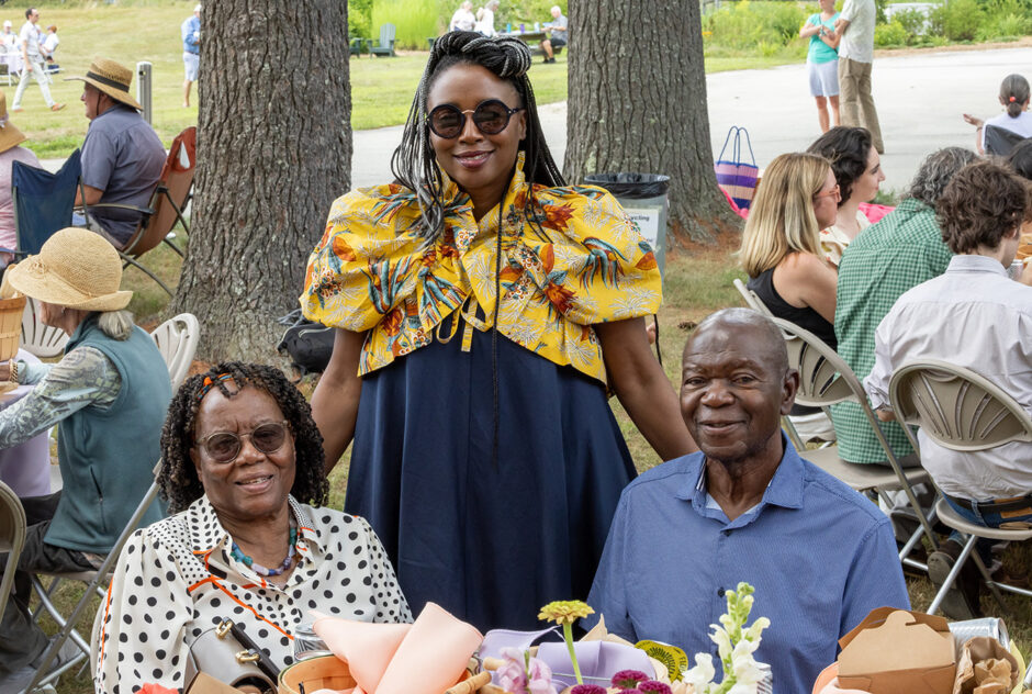 Three people smile warmly for a photo. Around them, people are gathered together for a picnic lunch