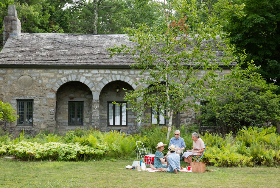 A small group gather together around a picnic on a lush lawn. Behind them, a stone building featuring arches create a bucolic backdrop