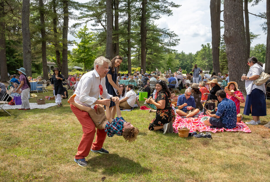 A man plays with his son while others are gathered around their picnic lunches. The scene is set in a small field