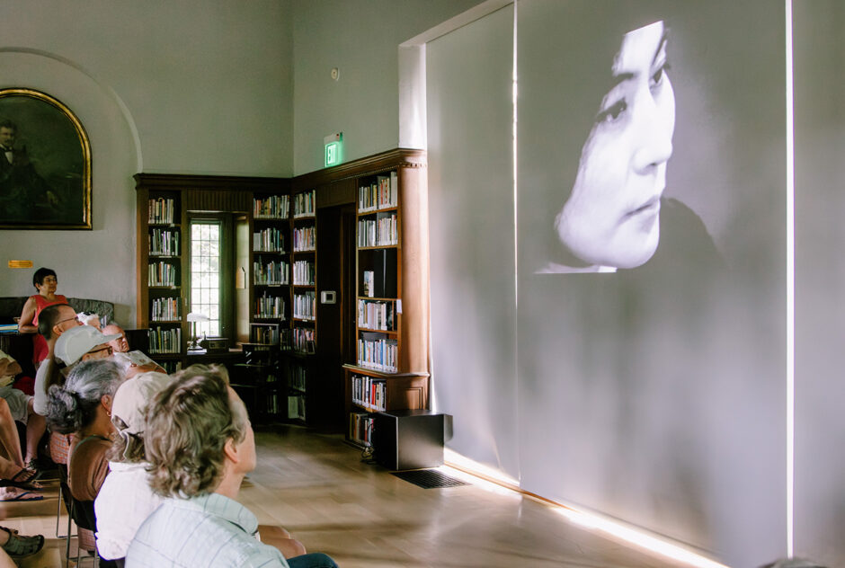 A small crowd gathers in a room to view films that are projected onto a large screen