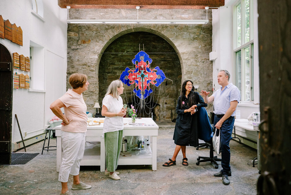 A group of four people gather inside an artist studio featuring a large stone arch and stone flooring. Behind them, a large colorful kite hangs on the wall