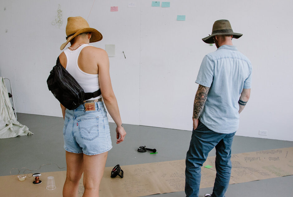 Two people stand in the center of a large room. They look toward the ground where an artist has set up a presentation of their work