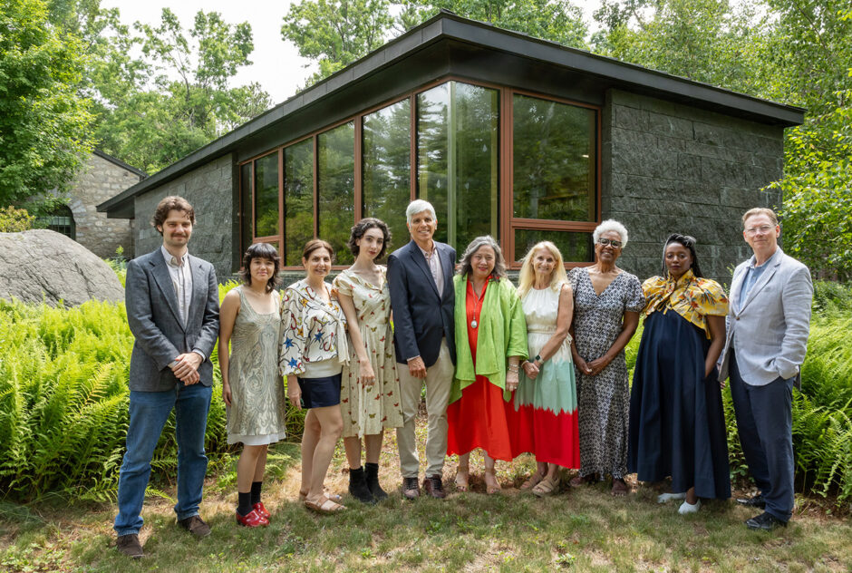 A large group of people stand in a row, posing for a picture. Behind them, a stone building with large windows is surrounded by lush ferns