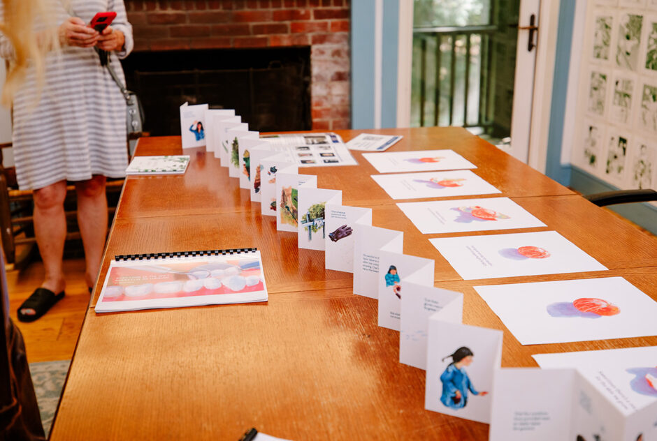 A long wooden table is adorned with artwork, one of which is an accordian folded piece of paper with drawings on it stretching several feet.