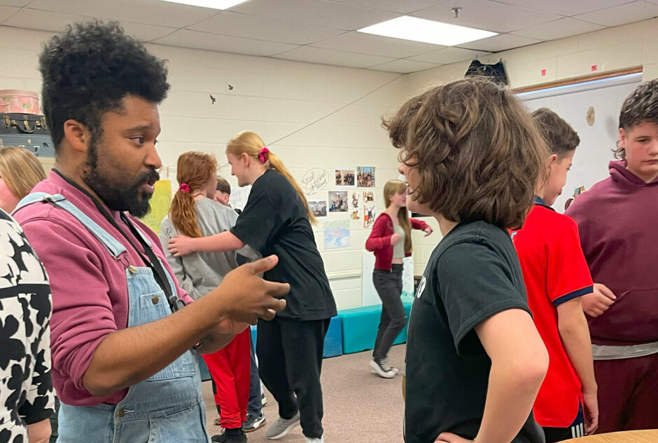 A man wearing overalls and a red long sleeve shirt stands face to face with a young student wearing a black t-shirt. While they have a discussions, other students work together in a similar manner throughout the room.