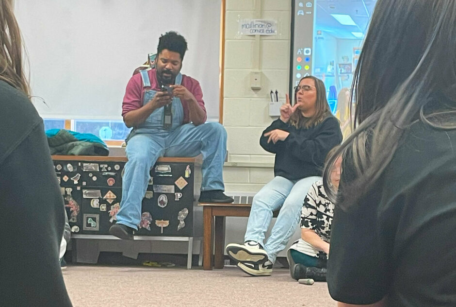 A man wearing overalls and a red long sleeve shirt sits at the front of a room, reading something from his phone. A woman, performing sign language translations, sits beside him.