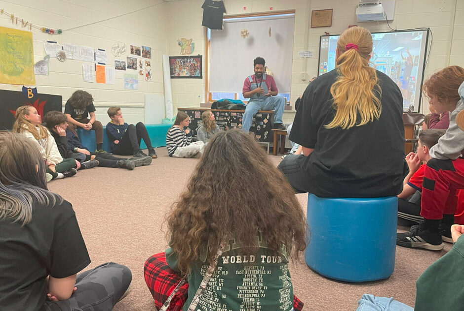 A man wearing overalls and a red long sleeve shirt sits at the front of a room. In front of him, students sit in a circle on the floor listening to him.