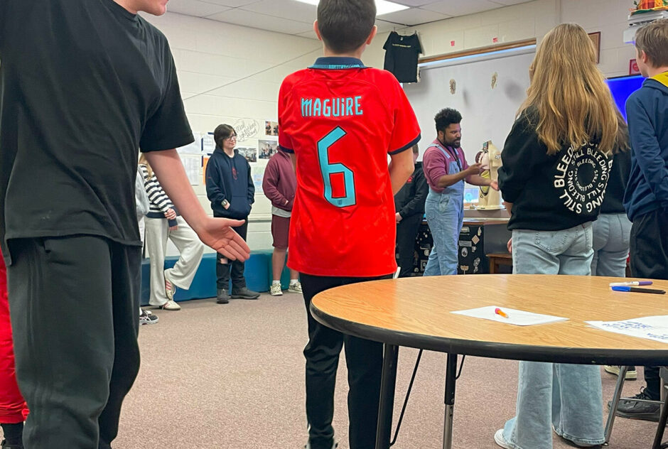 A man wearing overalls and a red long sleeve shirt stands at the front of a room. Surrounding him are students, also standing. The man gestures with his hands as he speaks.