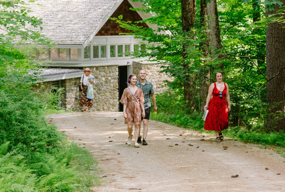 Four people make their way along a dirt road. Surrounding them is a lush green forest. I the distance, a stone building can be seen