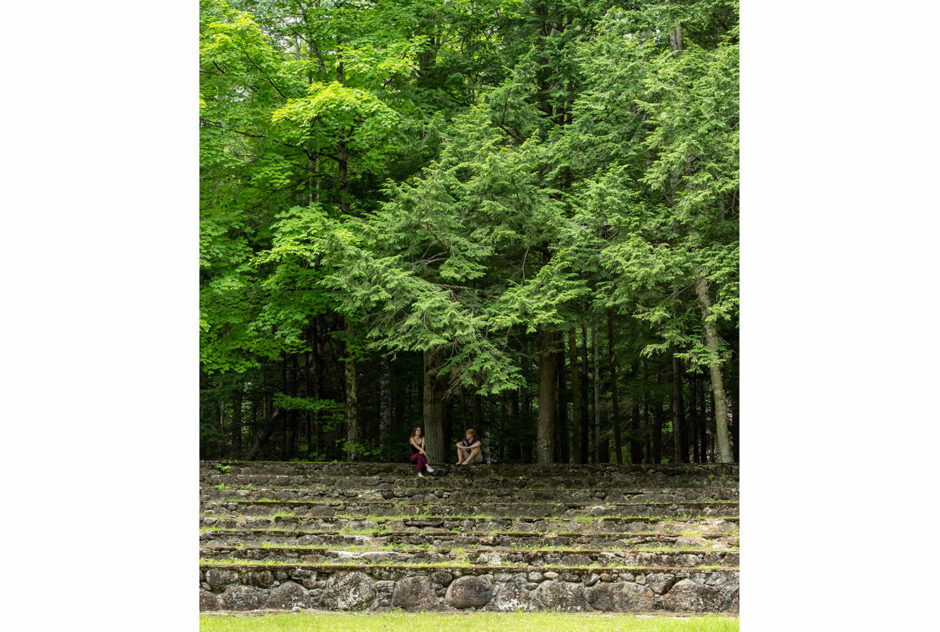 Two people sit atop a stone amphitheater. Lush green trees tower over the scene