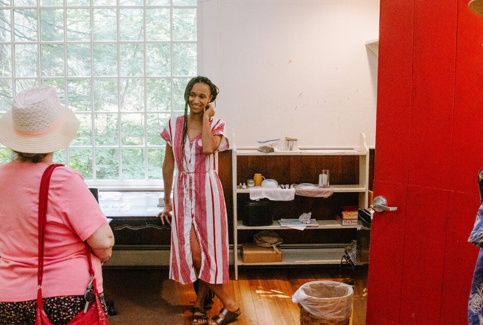 A woman in a red and white striped dress stands against a desk and speaks with another woman