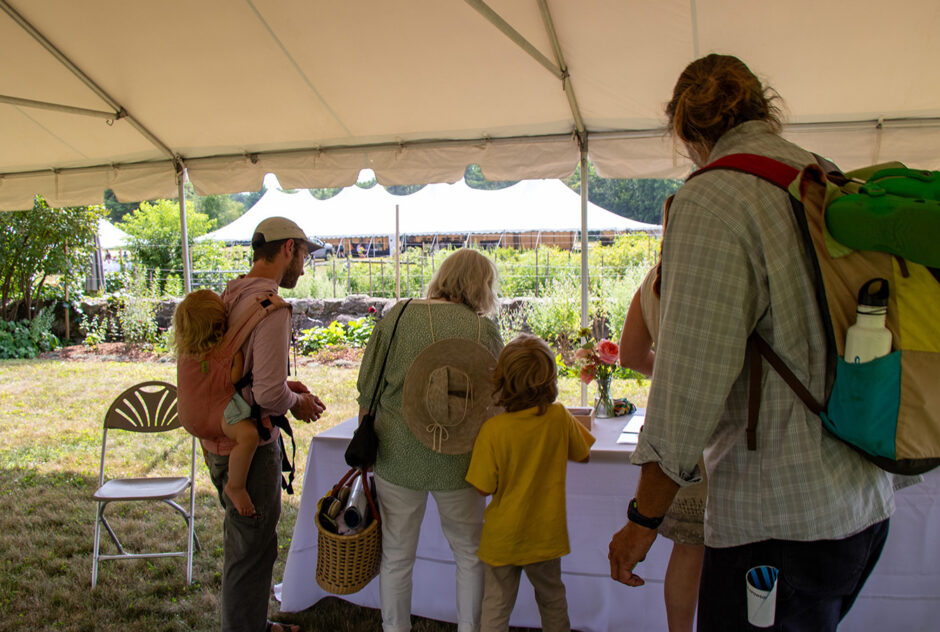 a small group gather under a white tent. They are gathering materials for an activity from a table