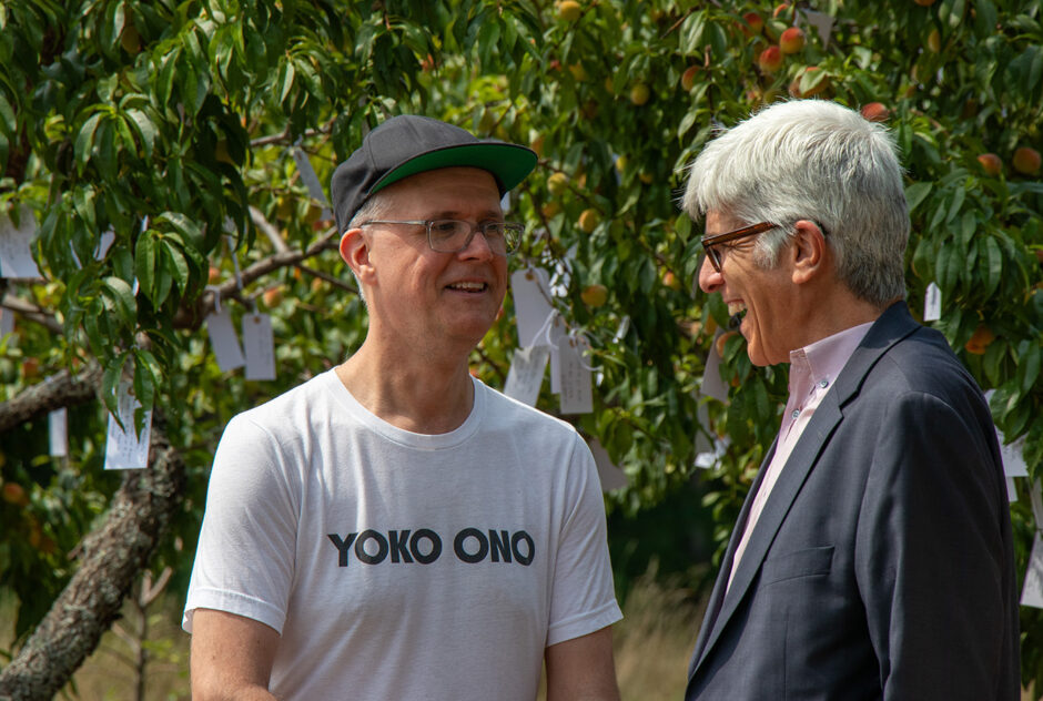 Two men stand together, chatting. The man on the right is laughing. Behind them is an orchard of peach trees