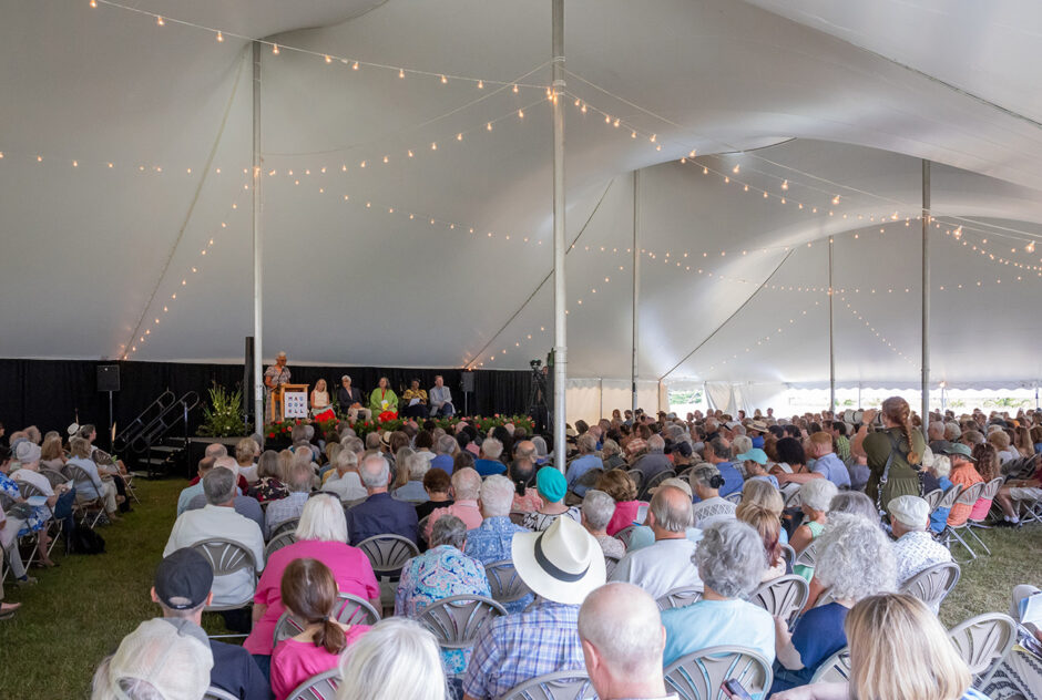 A large crowd sits in rows of chairs under a large white tent. They are facing the stage where a speaker is at the podium