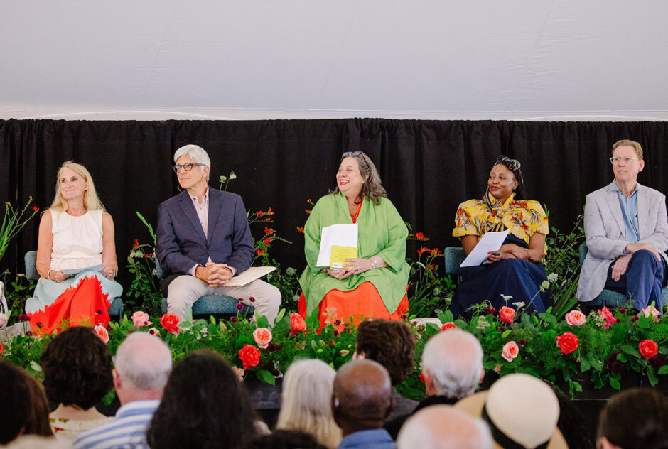 Five people sit on a stage, smiling and looking toward the speaker. In front of them, a crowd is gathered to listen.