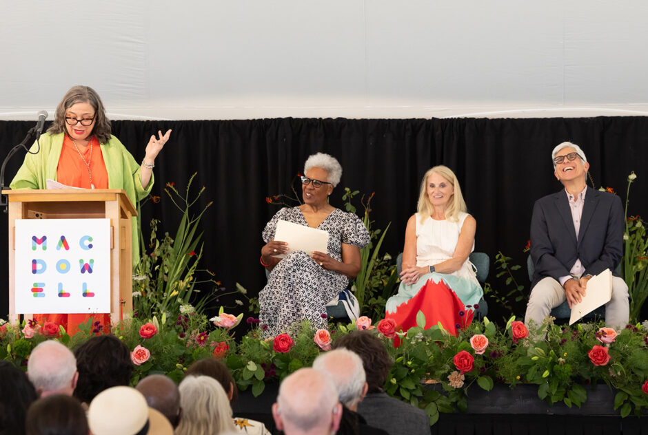 A woman stands at a podium and addresses a large crowd. Beside her on stage, the other speakers sit in a row of chairs