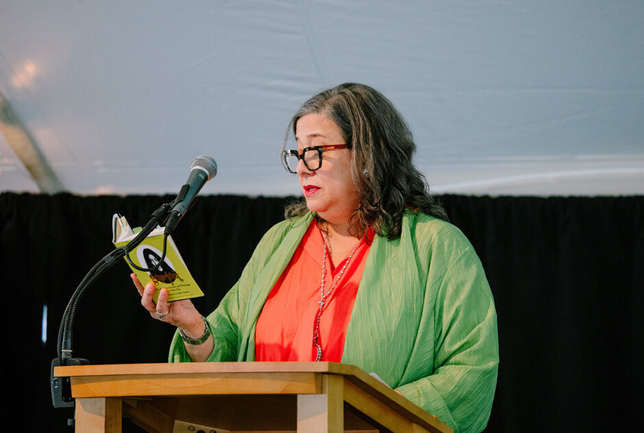 A woman wearing vibrant colors stands at a podium and reads from a small yellow book