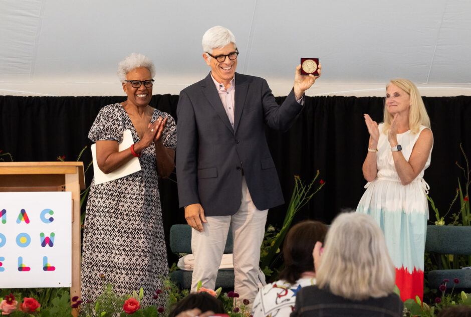 A man stands on stage and smiles widely as he holds up the Edward Macdowell Medal triumphantly. Other people on the stage smile toward him while they applaud