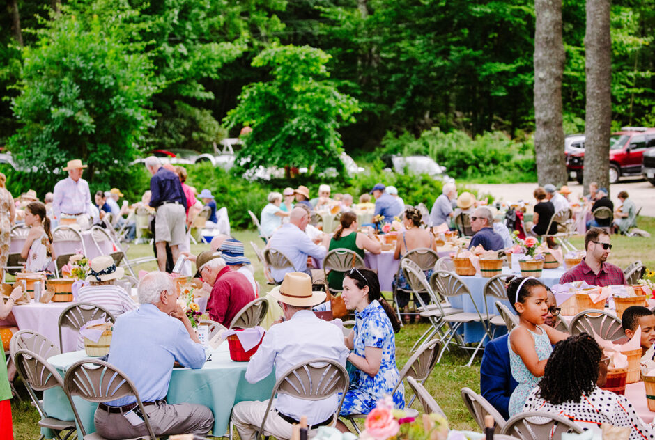 A small field is filled with guests who sit on picnic blankets and at tables, enjoying their lunch and one anothers company