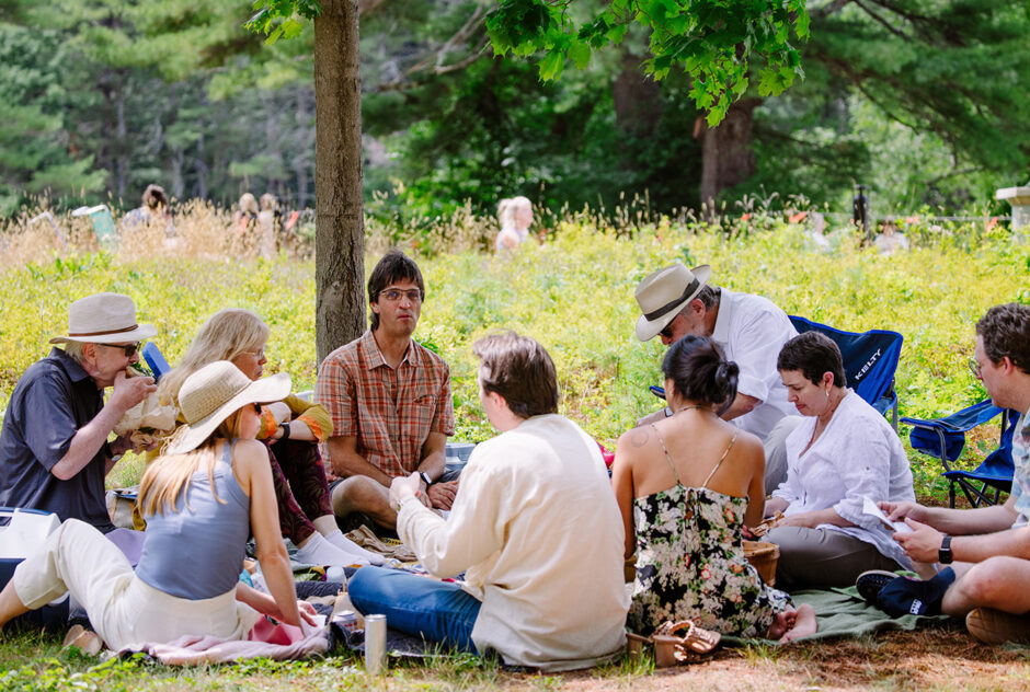 A group of people gather on a picnic blanket and enjoy their lunches and conversation. Behind them, a lush meadow glows in the sunlight