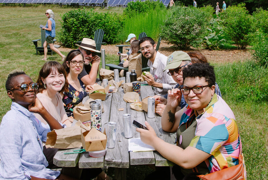 A group of people, smiling toward the camera, gather around a picnic table to eat thei lunch