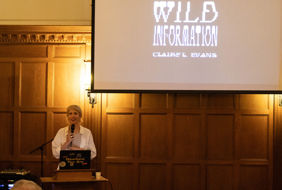 A woman stands at the front of a large, wood-paneled room. She speaks into a microphone from behind a podium. Behind her, a large projector screen hangs on the wall.