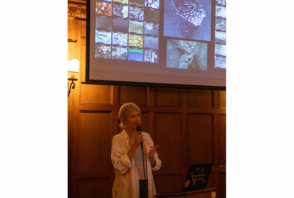 A woman stands at the front of a large, wood-paneled room. She speaks into a microphone from behind a podium. Behind her, a large projector screen hangs on the wall.