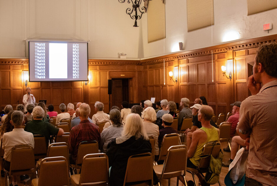 A woman stands at the front of a large, wood-paneled room. She speaks into a microphone from behind a podium. Behind her, a large projector screen hangs on the wall. Rows of chairs are filled by audience members who listen carefully to the presentation.