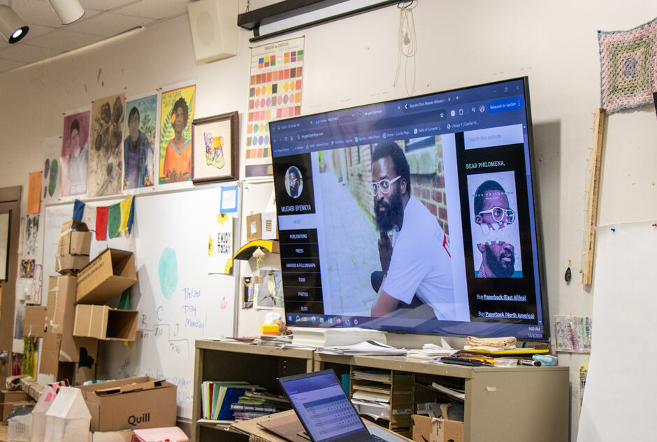 An art classroom. The walls are covered in artworks and papers. A large TV screen displays an image of a man.