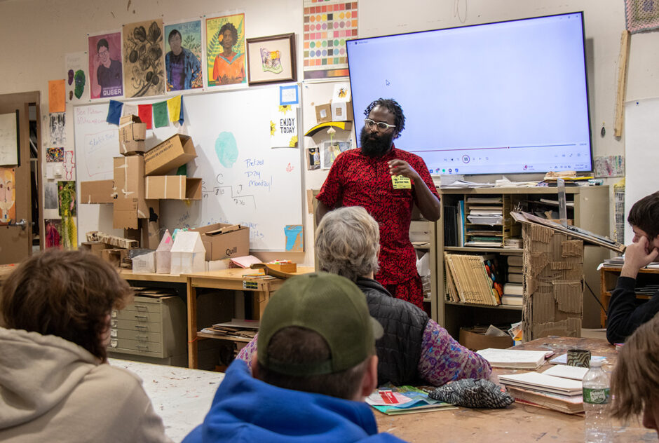 A person wearing a bright red outfit stands at the front of an art classroom, speaking and gesturing with their hand. The walls of the classroom are covered in artwork and papers. Behind the person is a large TV screen.