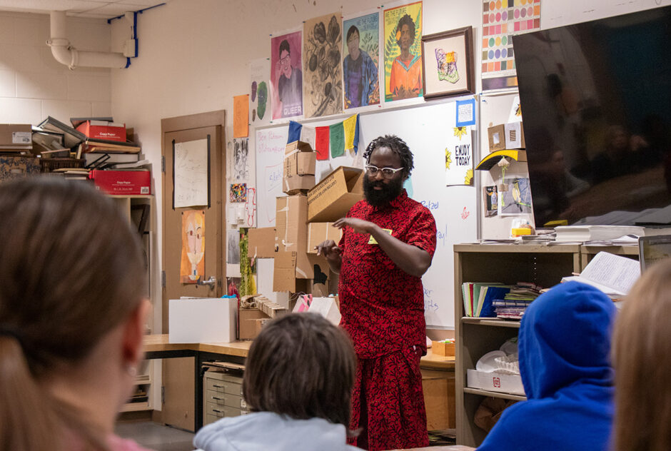 A person wearing a bright red outfit stands at the front of an art classroom, speaking and gesturing with their hand. The walls of the classroom are covered in artwork and papers. Behind the person is a large TV screen