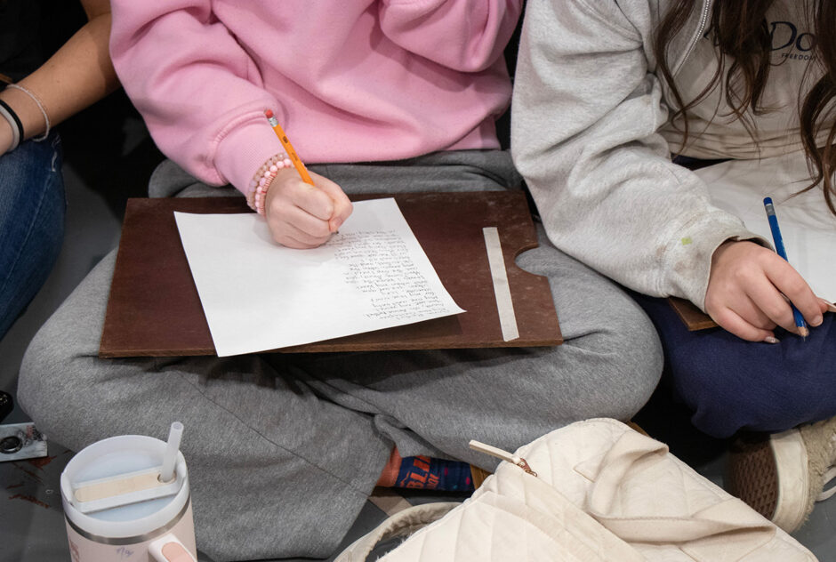 A close-up shot of two students who are sitting cross-legged on the floor. They are writing poems on paper.