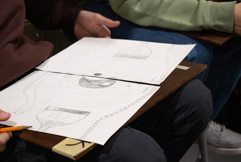 A student holds two drawings in his lap. He and a friend created them together. The two pages, when placed next to one another, create one larger artwork