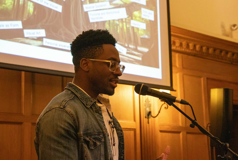 A man stand at a podium in a warmly lit room. Behind his, a projector screen displays a collage of words.