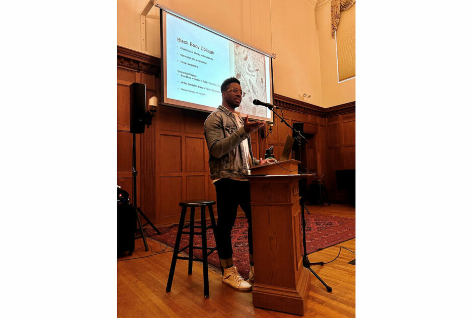 A man stand at a podium in a warmly lit room. Behind his, a projector screen displays a slide with information about the speaker's musical process