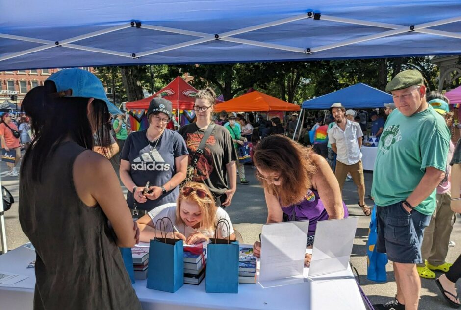 People sign their names on a sheet of paper at a table under a small tent during a street festival