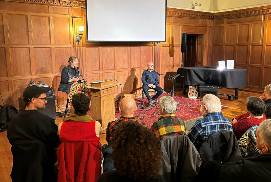 A small crowd, sitting in rows of chairs, face the front of a large, warmly lit room. Two artists sit at the front and are presenting their art practices