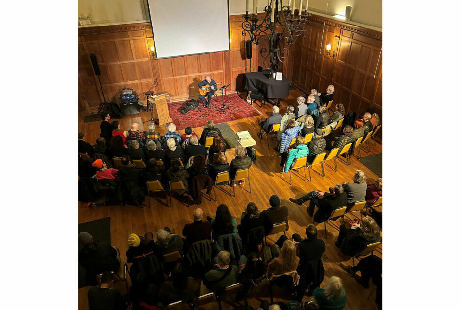 A crowd, sitting in rows of chairs, face the front of a large, warmly lit room. An artists sits at the front and plays guitar