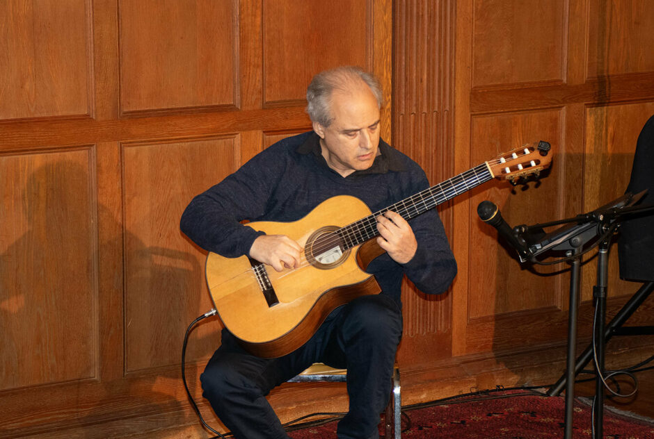 A man sits in a warmly lit, wood paneled room. He is playing the guitar