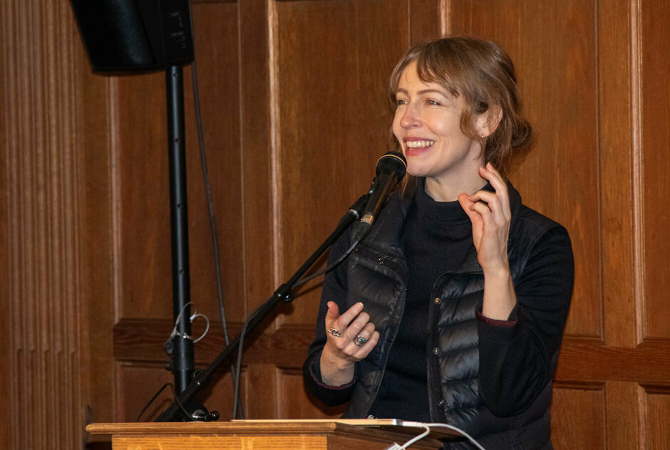 A woman stands at a podium in a wood paneled room. She is gesturing with her hands and smiling.