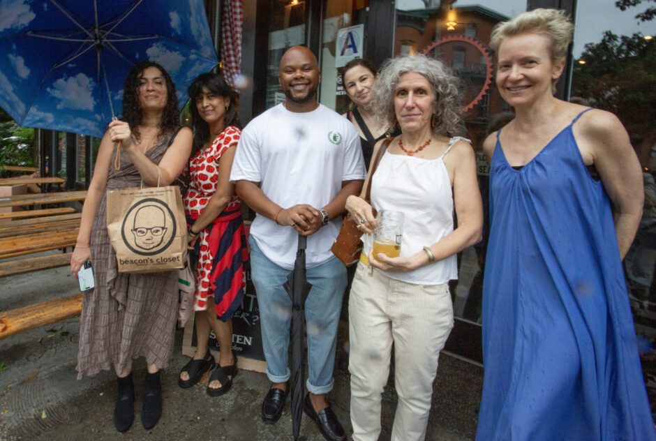 Six people in summer clothing, some holding umbrellas, pose together for a photo on the sidewalk outside a New York bar.