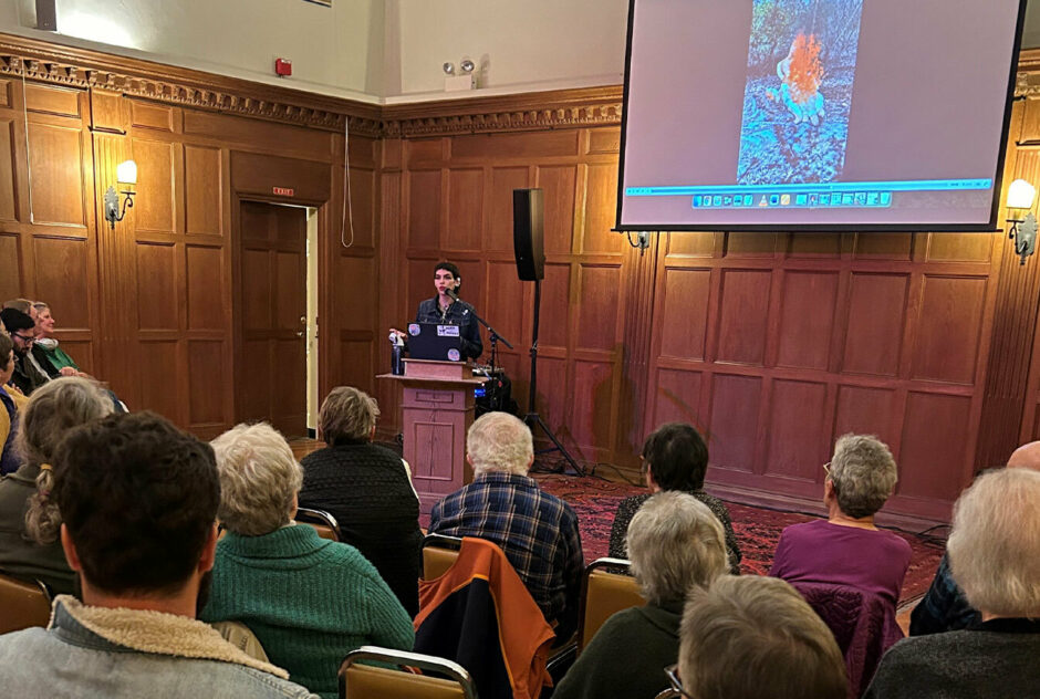 A person stands at a podium at the front of a large room, filled with people. They are referencing images projected onto a large screen as they deliver their presentation