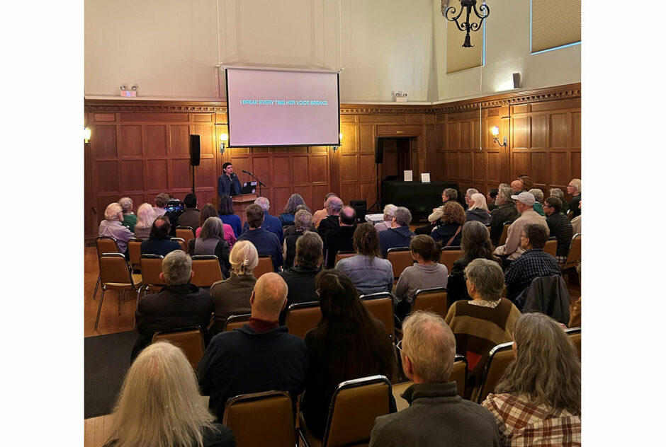 A person stands at a podium at the front of a large room, filled with people. They are referencing images projected onto a large screen as they deliver their presentation