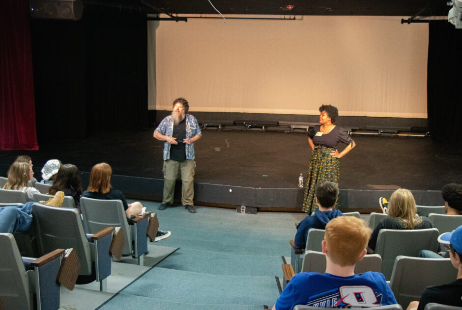 Two adults stand at the front of a small auditorium, just in front of the stage. They are introducing the activities for the day. Students, sitting in the rows of chairs, face them.