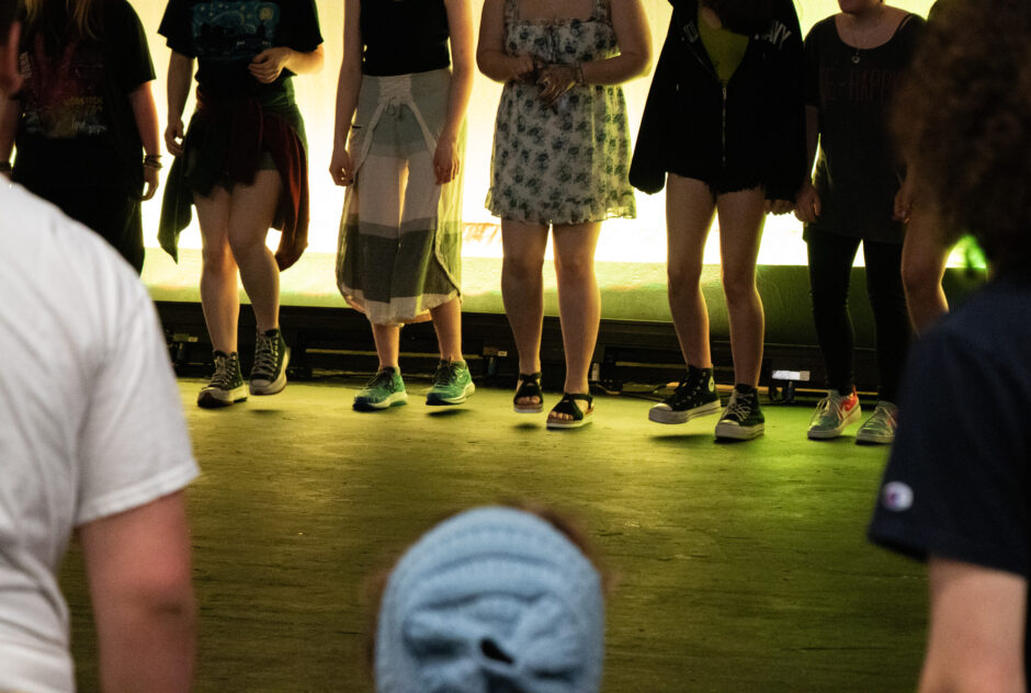 A row of students stand on a stage, a yellow light illuminating the scene. You can see them from the chest down, as they stomp their feet in unison.