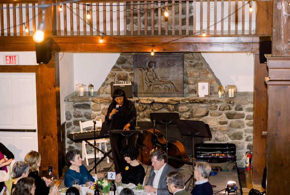 A woman on stage in front of a fireplace speaks into a microphone