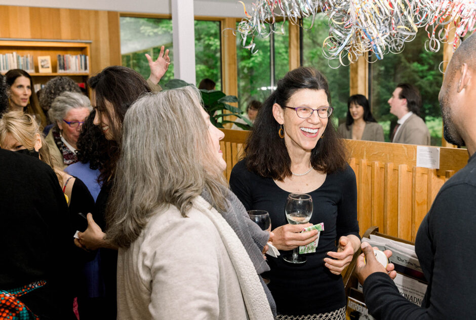 A woman smiles and laughs while holding a drink and speaking with two people whose backs face the camera