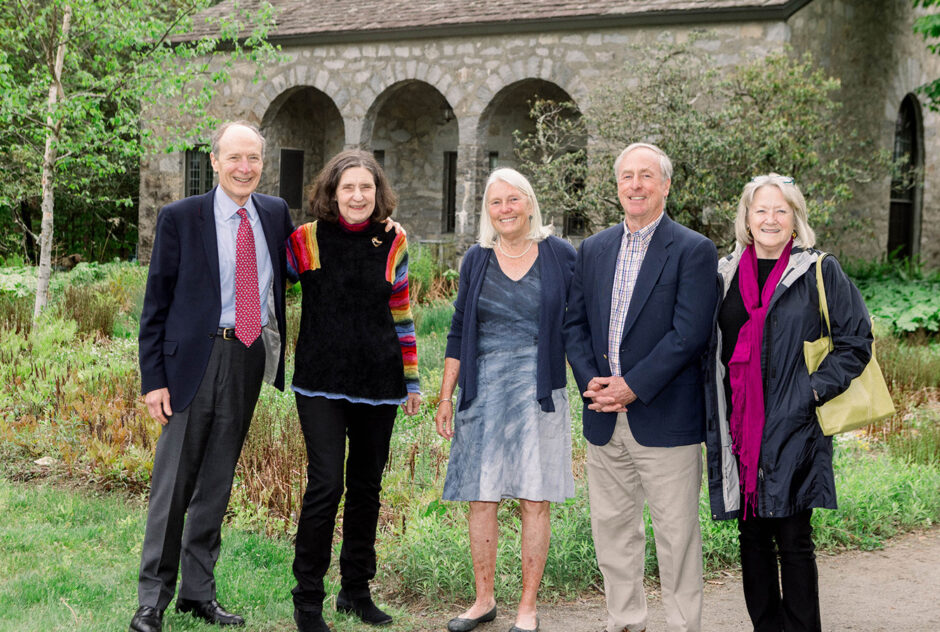 A group of five older adults wearing suits and dresses pose for the camera in front of a stone building and a grass lawn