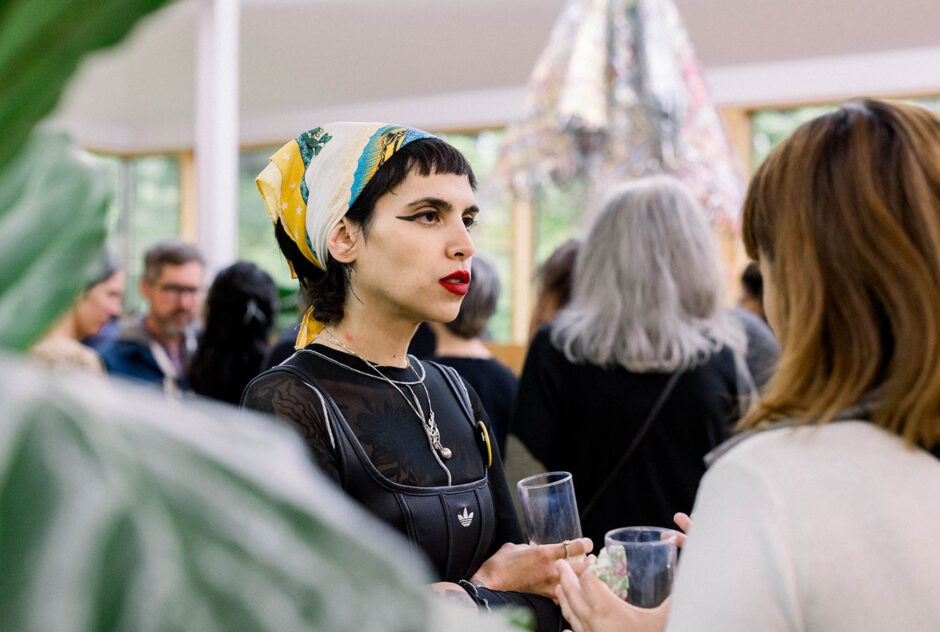 A woman in a colorful bandana is in conversation with someone who's back is to the camera. A plant leave blocks part of the camera view