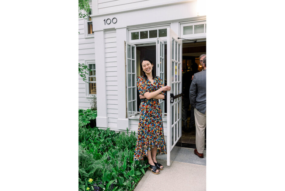 A woman in a colorful dress smiles and opens a door to a white building